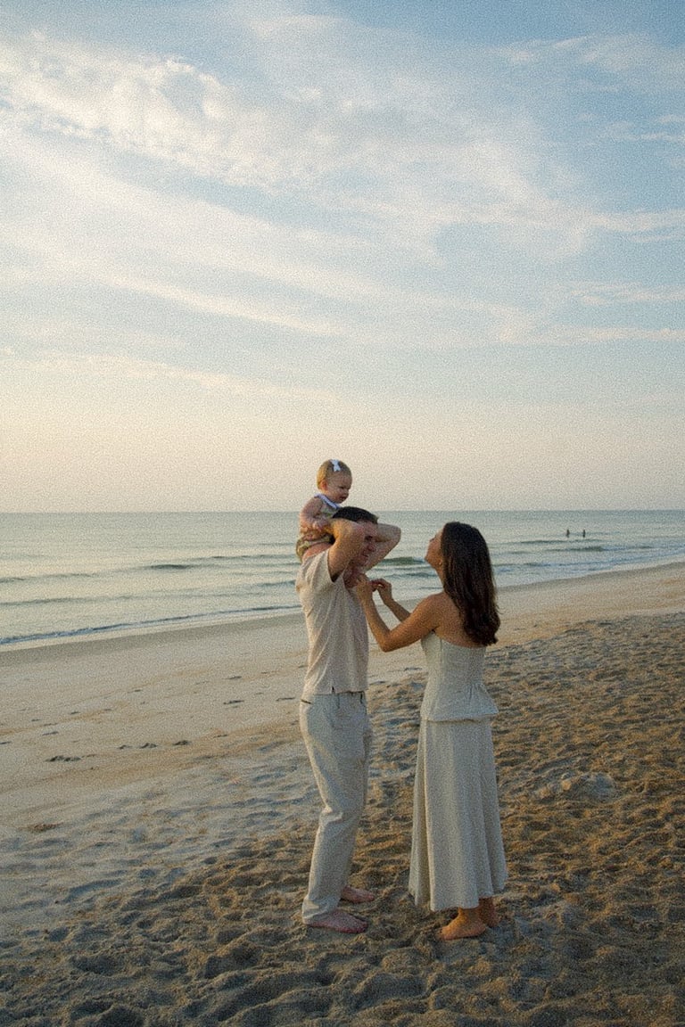 Family of three in white clothing playing on a sandy beach at sunset with calm ocean waters in the background