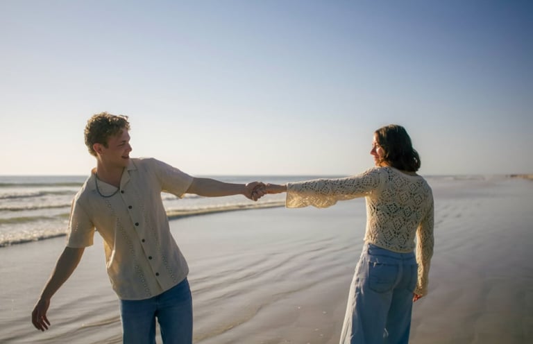 Two people holding hands walking along a sandy beach at golden hour with ocean waves in the background