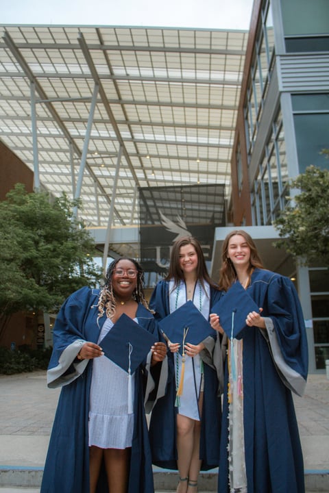 Three female graduates in blue robes holding diplomas stand together under a modern glass atrium building