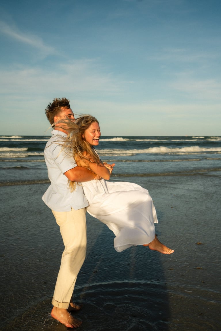Man in light clothing carrying woman in white dress on beach with ocean waves in background