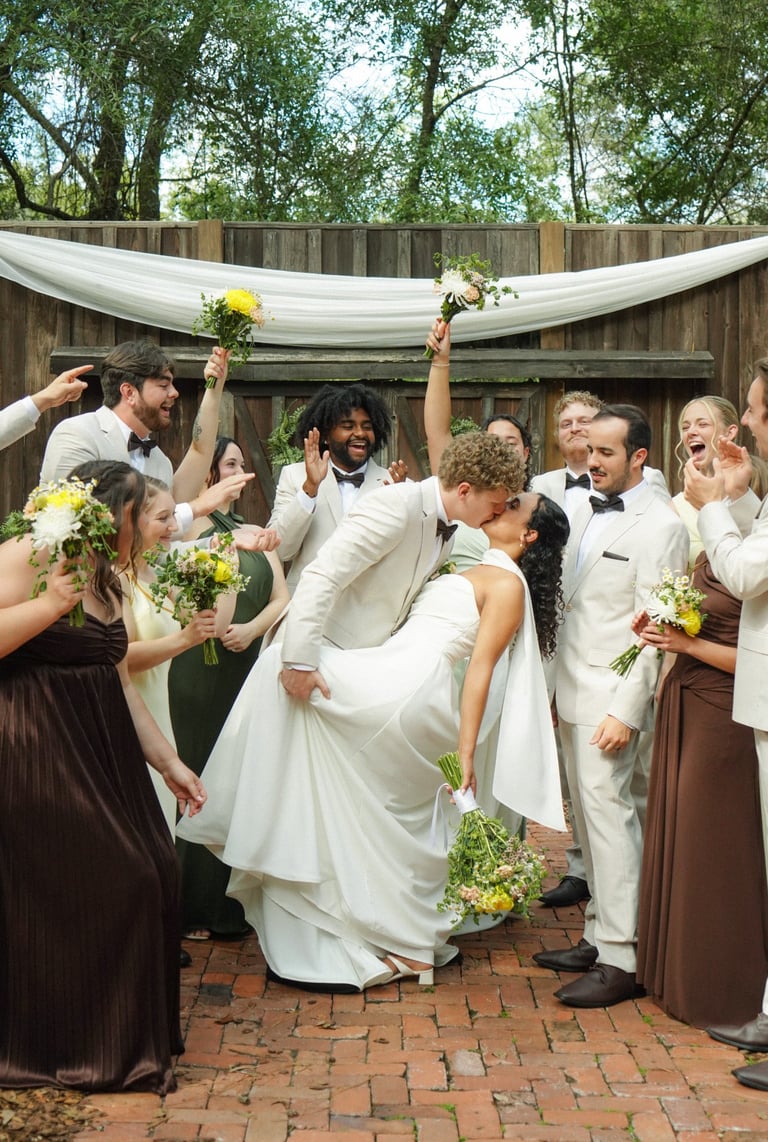 Groom lifts bride in celebration surrounded by wedding party holding yellow flowers in backyard garden ceremony