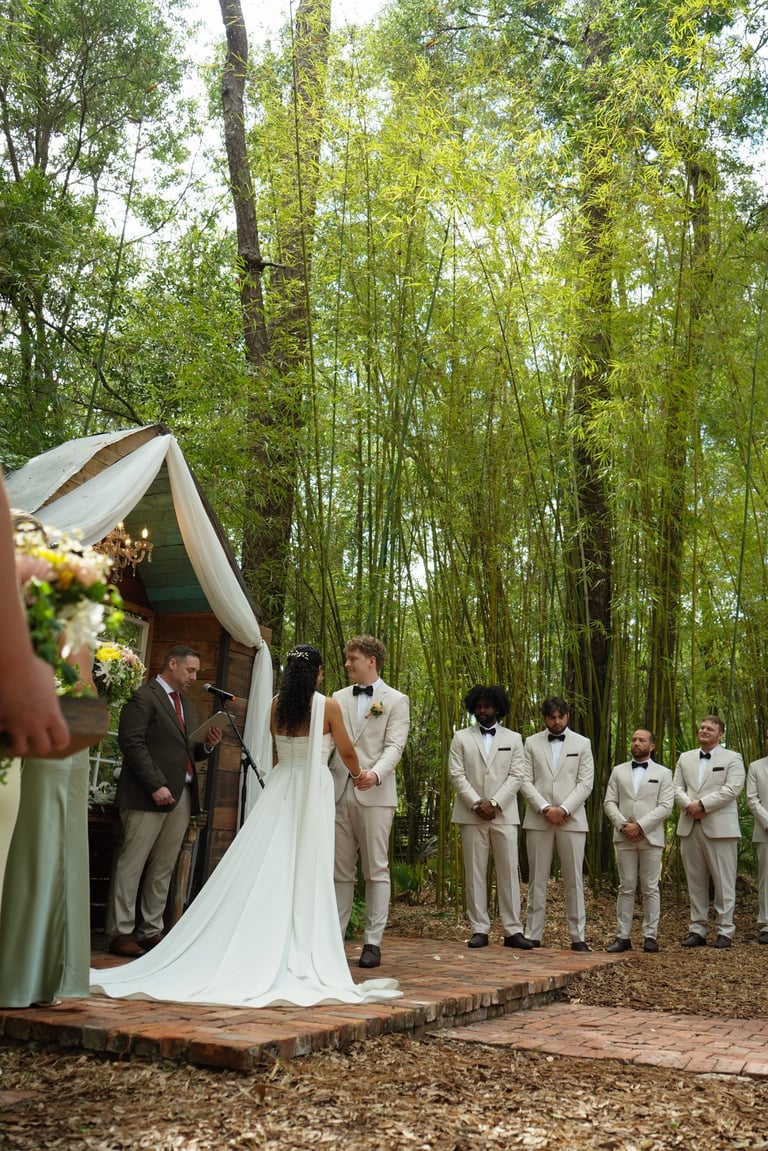 Wedding ceremony in a bamboo forest with bride in white gown and groom in beige suit exchanging vows under a draped arch