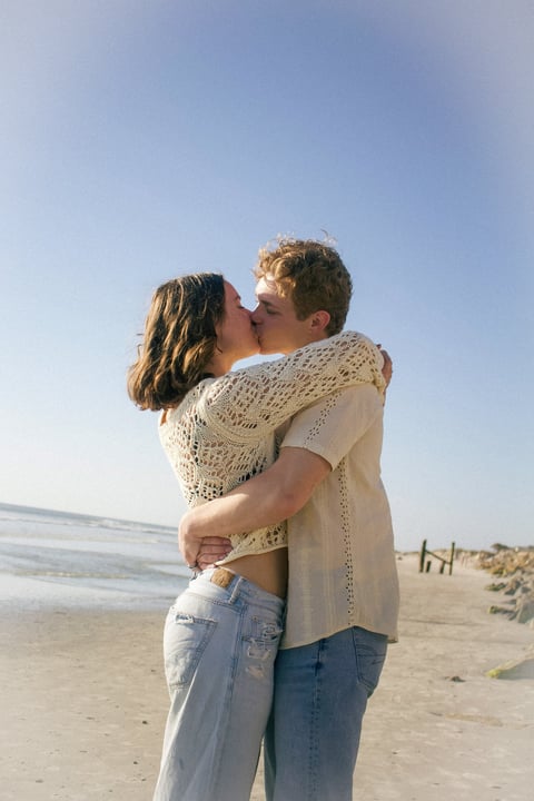 Couple kissing on a beach in casual clothing with the ocean and clear sky in the background