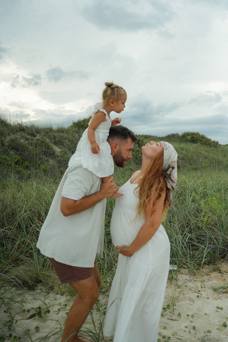 Family of three in white clothing at the beach with sand dunes and grassy area in background