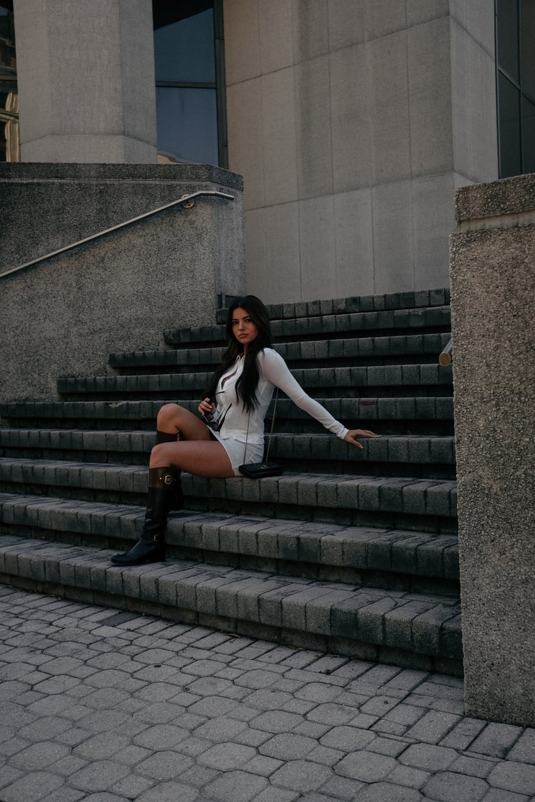 Woman in white top and boots sitting on concrete stairs in front of modern building