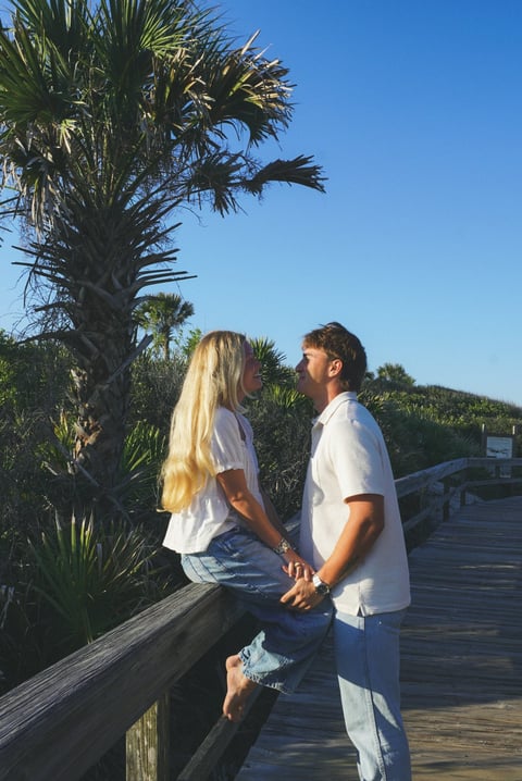 Couple holding hands on a wooden boardwalk surrounded by palm trees and tropical vegetation under clear blue sky