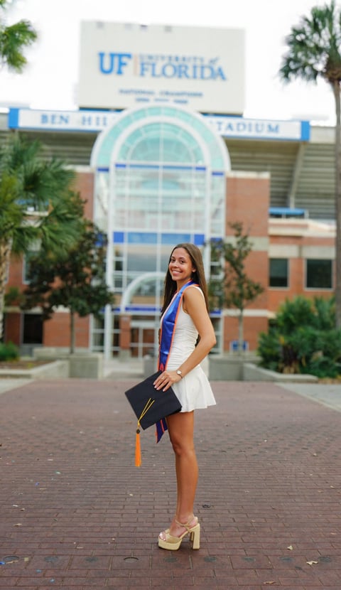 Woman in graduation attire holding diploma in front of UF Florida stadium building with palm trees