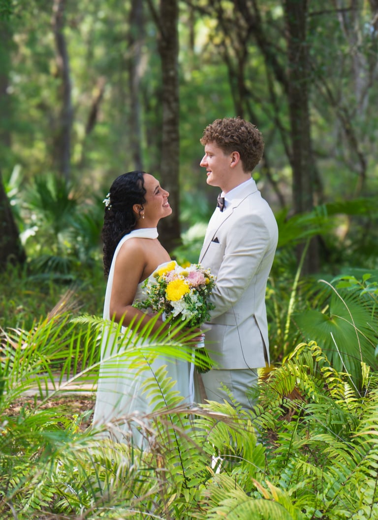 Bride and groom stand together in a lush forest setting, holding a yellow and white bouquet