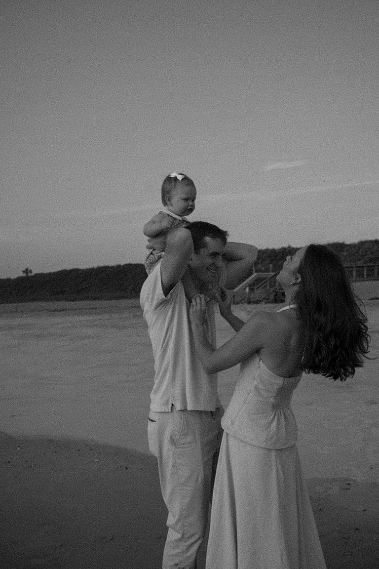 Black and white photo of a couple holding their young child on a beach, with hills in the background