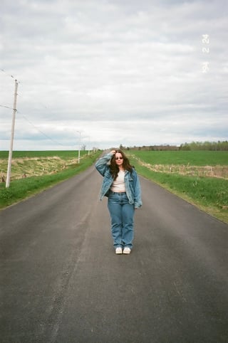 Woman in denim jacket standing on a rural road with green fields on either side under cloudy sky