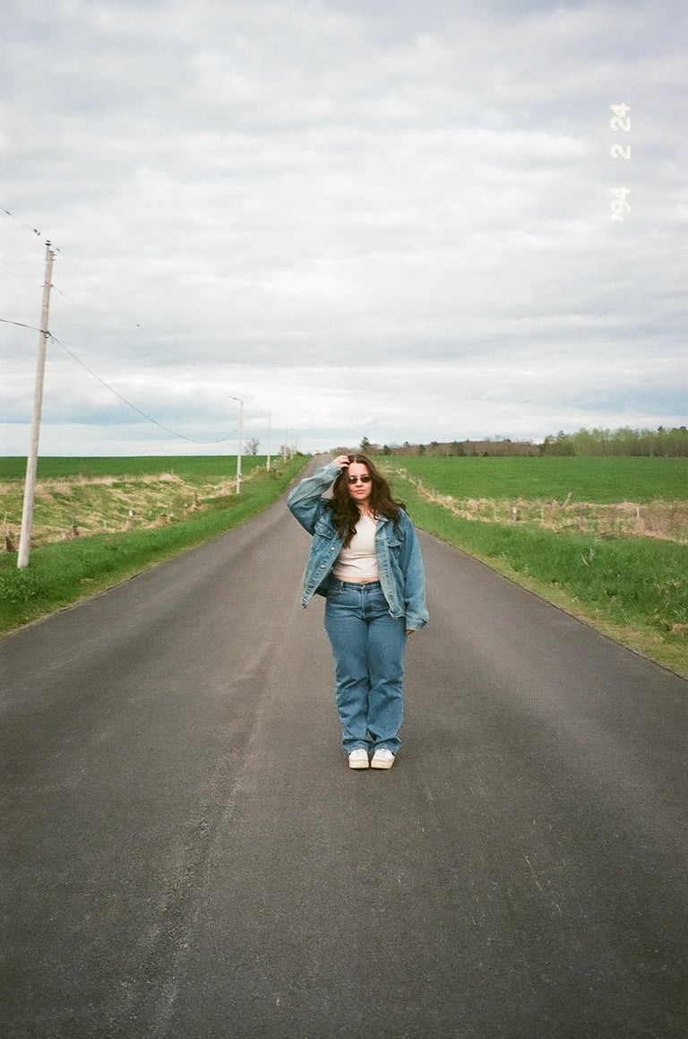 Woman in denim jacket standing on a rural road with green fields on either side under cloudy sky