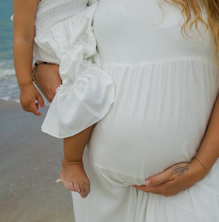 Pregnant woman in white dress holding baby bump at the beach