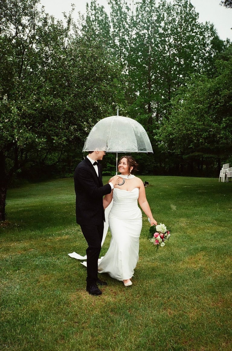 Bride and groom in white dress and black suit standing under clear umbrella on grassy lawn with tree canopy in background