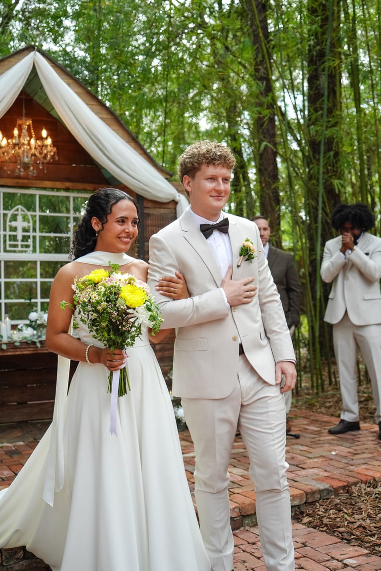 Bride and groom walk together outside wedding venue in forest. She wears white dress holding yellow bouquet, he wears cream suit with bow tie.