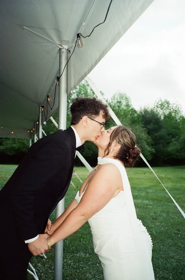 Bride and groom kissing under a tent in an outdoor garden setting with green grass and trees in the background