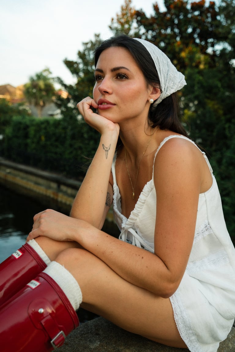 Woman in white headscarf and tank top sitting outdoors with hand to face, wearing white container, trees in background