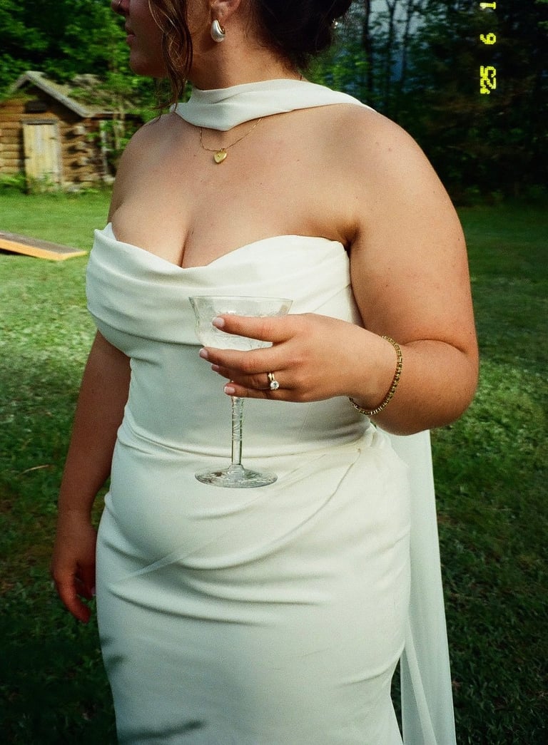 Woman in white off-shoulder dress holding a champagne glass at outdoor garden event, viewed from behind