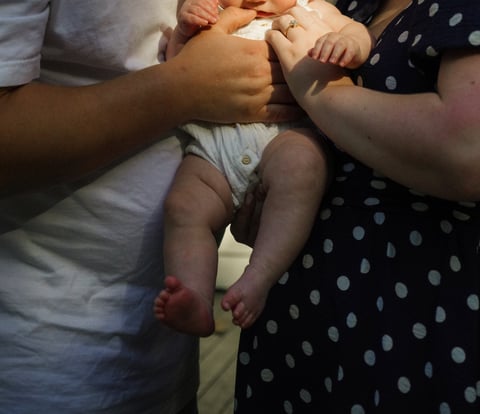 Two adults hold an infants legs and torso, displaying a bonding moment with the baby wearing a white diaper