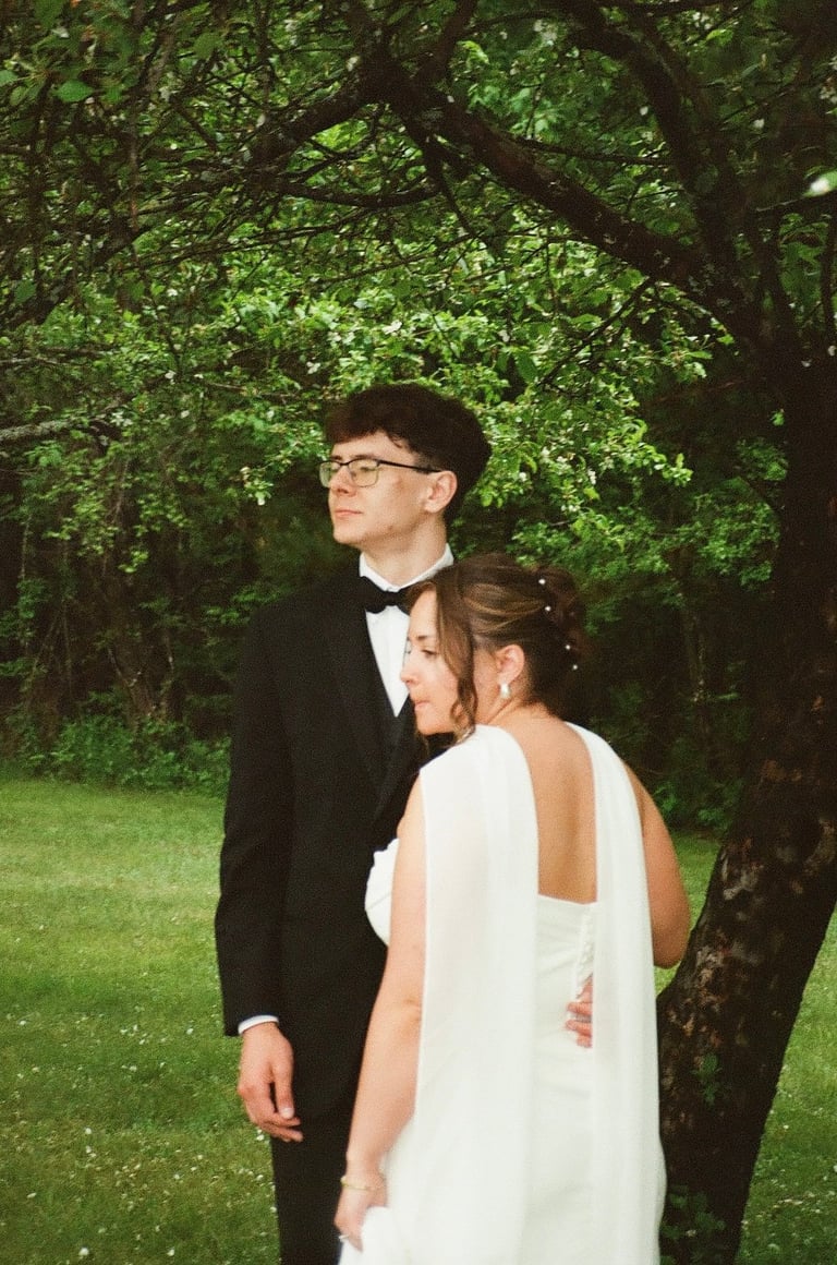 Couple in formal wedding attire standing together under large green trees in a garden setting