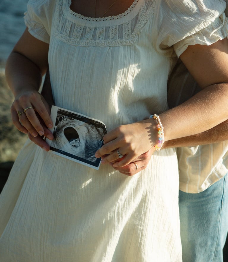 Couple holding ultrasound image, woman in white dress and man in jeans by ocean
