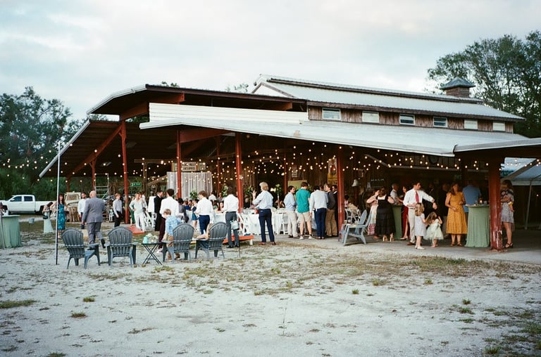 Outdoor wedding reception venue with guests gathered under a covered wooden structure decorated with string lights at dusk