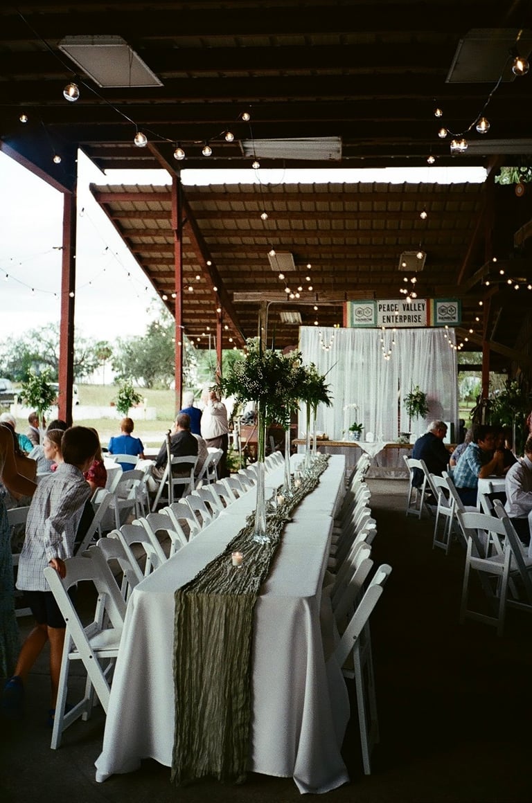 Indoor wedding reception venue with white chairs, draped tables, hanging lights, and guests seated in rustic barn setting