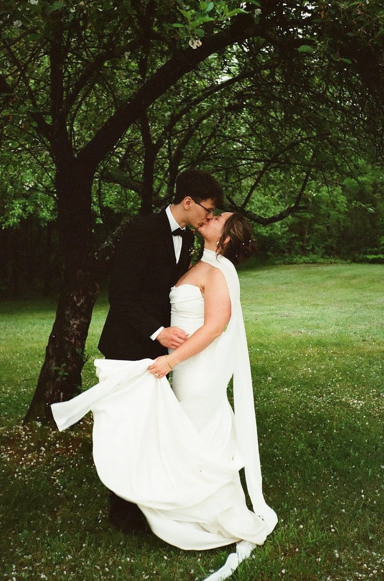 Newlyweds kissing outdoors under a tree canopy, groom in dark tuxedo and bride in white flowing dress on grassy lawn