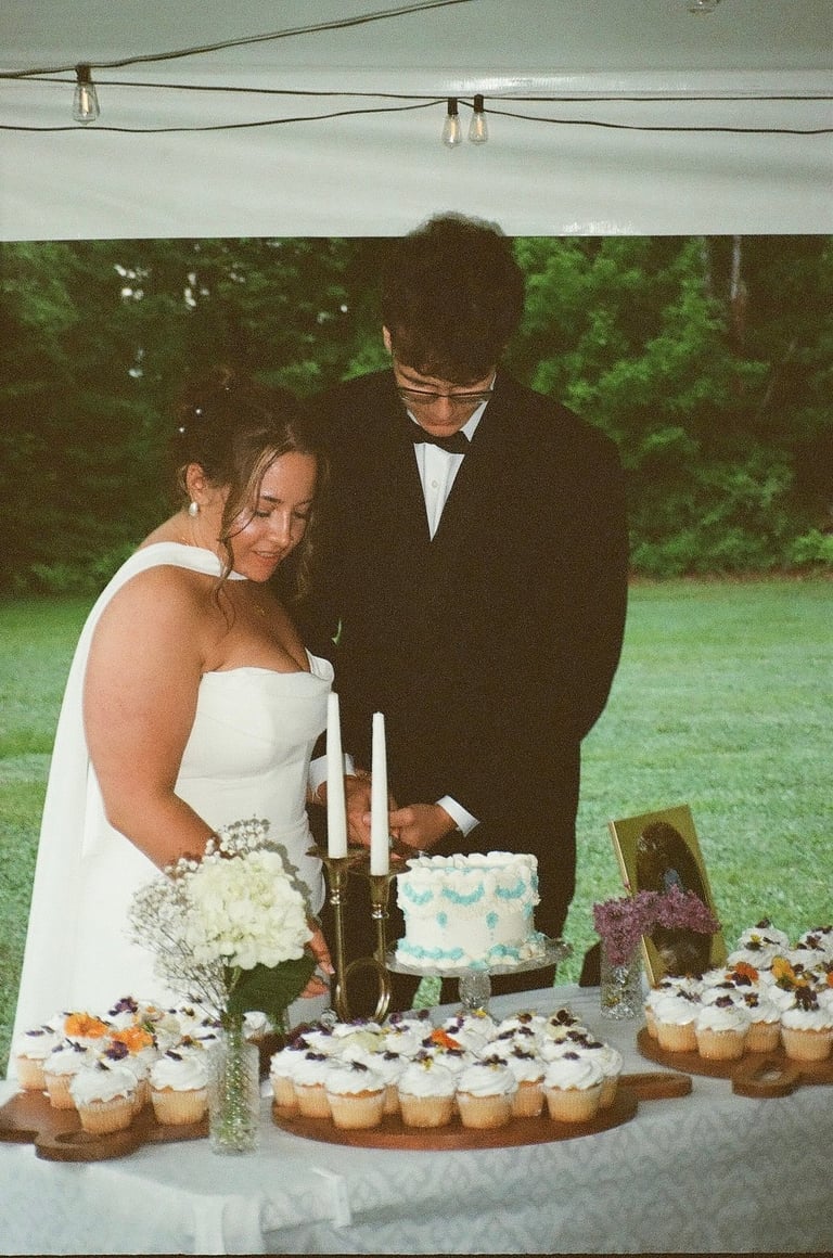 Bride and groom cutting wedding cake outdoors under a tent with cupcakes and flowers displayed on the table