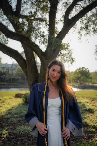 Woman in blue cardigan and white dress standing under large tree by water at sunset