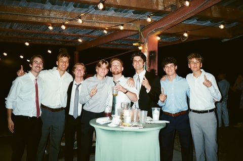 Group of eight men in formal attire posing together at an indoor event with a green table and overhead lighting
