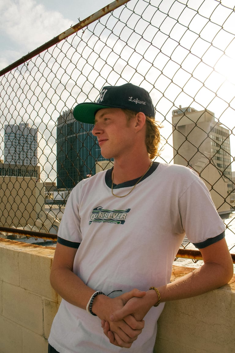 Young man wearing green cap and white graphic t-shirt posing on rooftop with city skyline and chain-link fence behind him