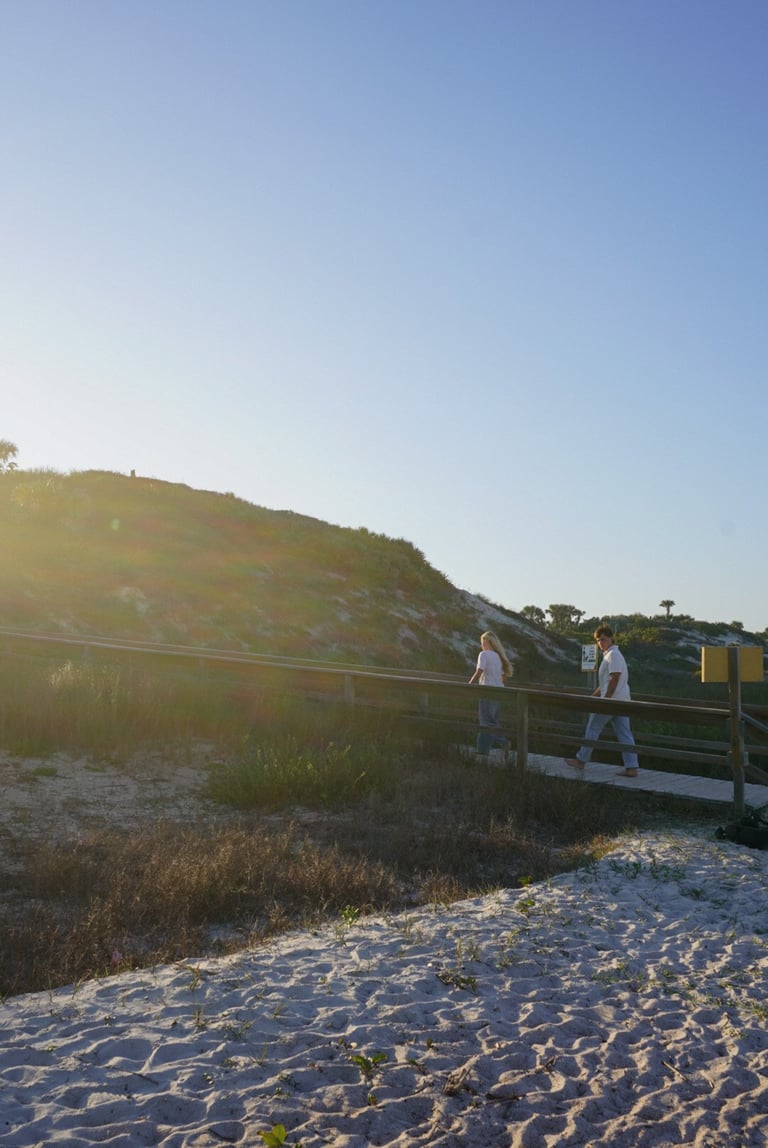 Two people walking on a wooden boardwalk across sand with dune grass and grassy hills in the background under a clear blue sky