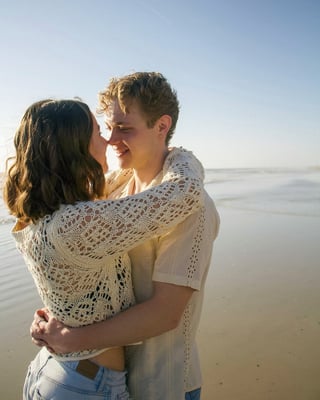 A young couple embracing on a beach at sunset, both wearing white knit sweaters and smiling together with the ocean behind them