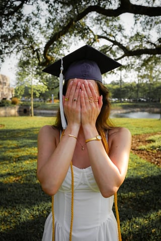 Graduate in cap and gown covering face with hands in celebration, standing outdoors on a campus lawn