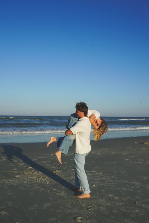 Man in white shirt lifting and holding a woman in his arms on a sandy beach with ocean waves in the background