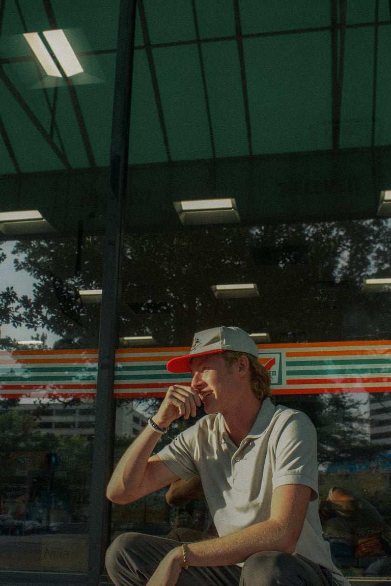 Man wearing red cap and white shirt sitting indoors with hand near face, glass panels and colorful striped background visible
