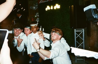 Four men in casual clothing posing playfully at a nightclub or bar with neon lights and green ivy wall backdrop