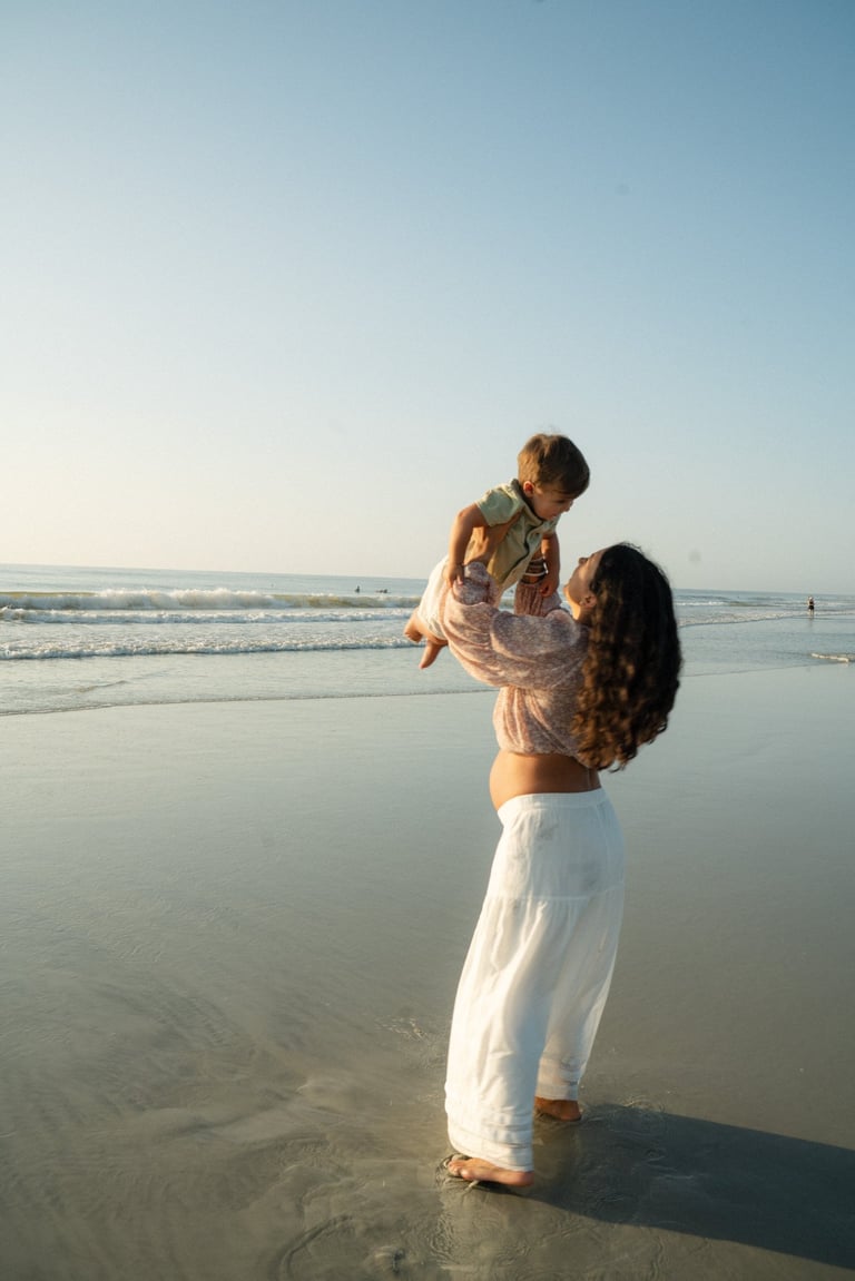 Woman holding a child up in the air on a beach with ocean waves in the background at sunset