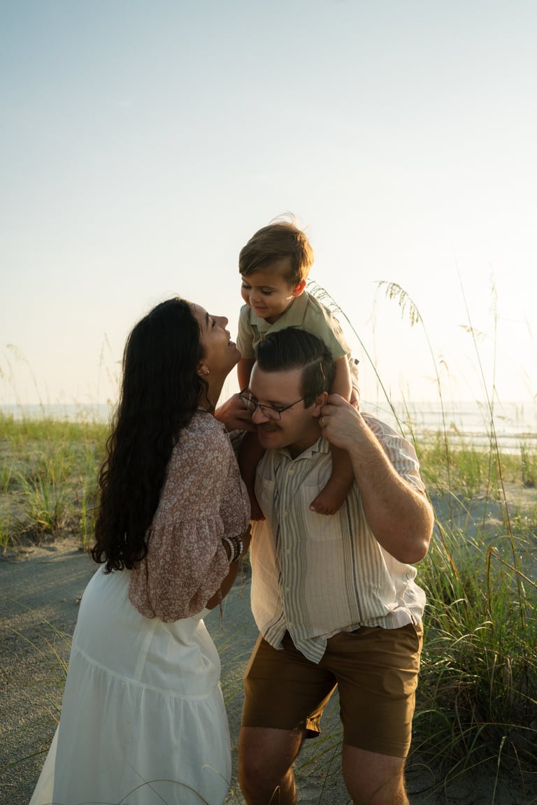 Family of three enjoying a beach walk at sunset, with father holding young son on shoulders while mother stands beside them