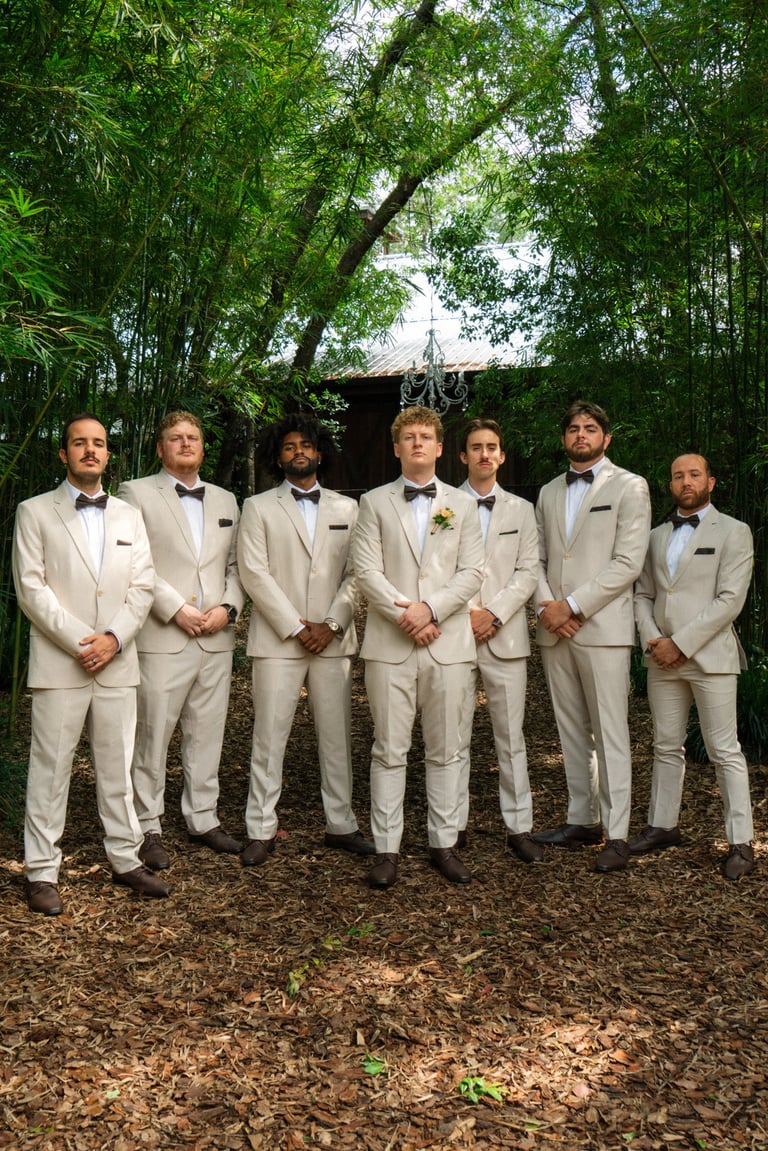 Seven groomsmen in beige suits and bow ties standing in a line under lush green bamboo trees