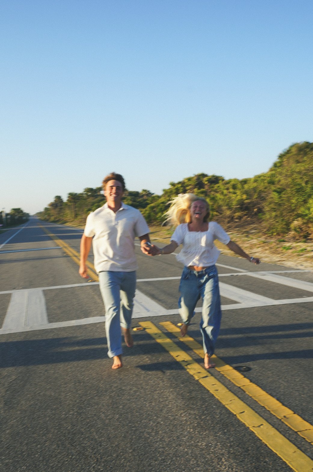 Couple jogging down an empty highway with yellow center lines, surrounded by green vegetation under clear blue sky