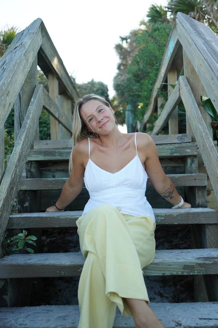 Woman in white tank top and yellow skirt sitting on wooden beach stairs, smiling at camera with palm trees in background