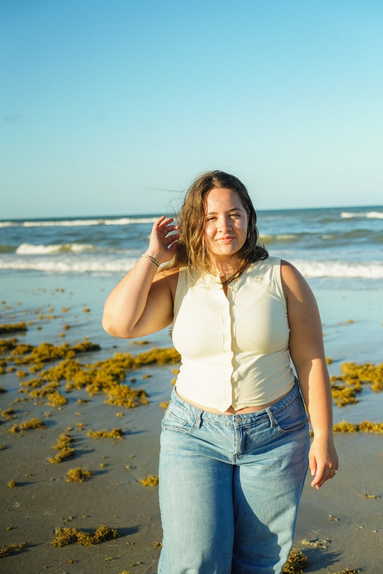 Woman in white top and jeans standing on beach with seaweed and ocean waves in background