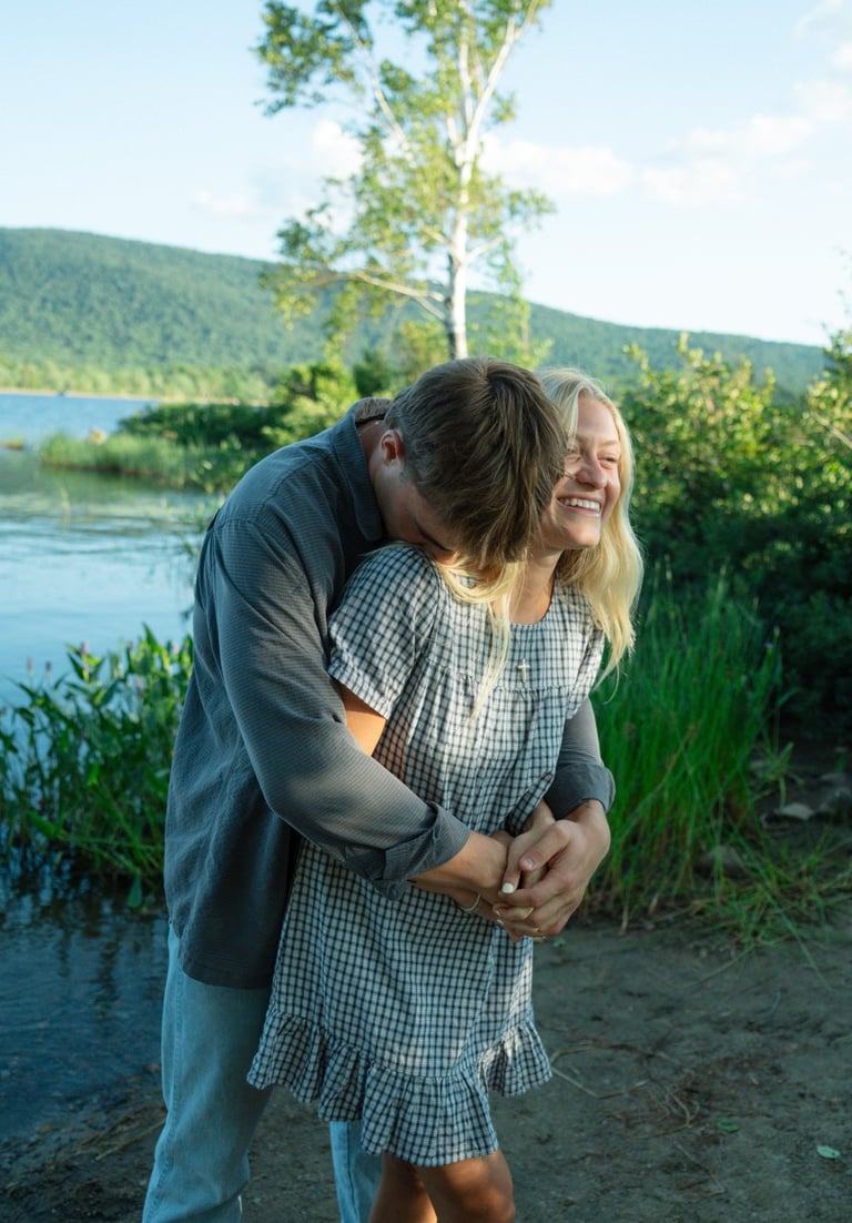 A man embraces a smiling woman from behind by a calm lake, with forested mountains and green vegetation in the background