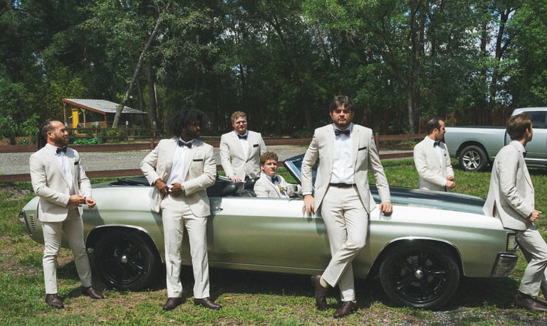 Groom and groomsmen in cream suits posing with a vintage green and white car at an outdoor wedding venue