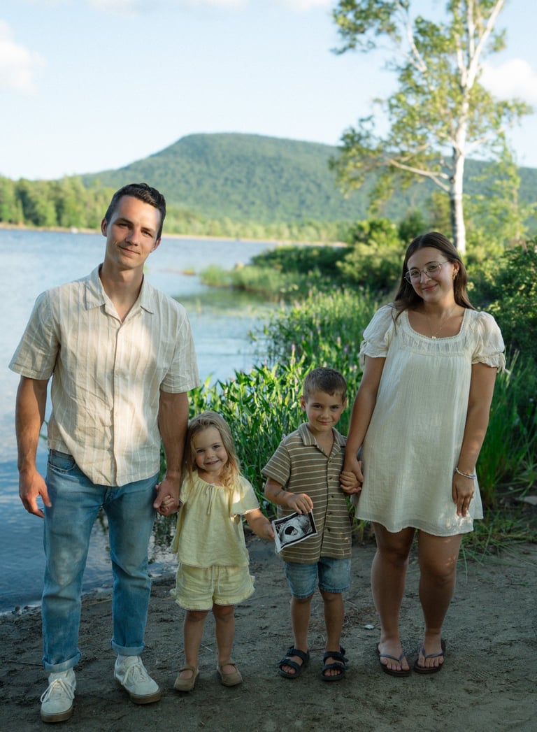 Family of four standing on sandy beach near lake with forested mountain in background