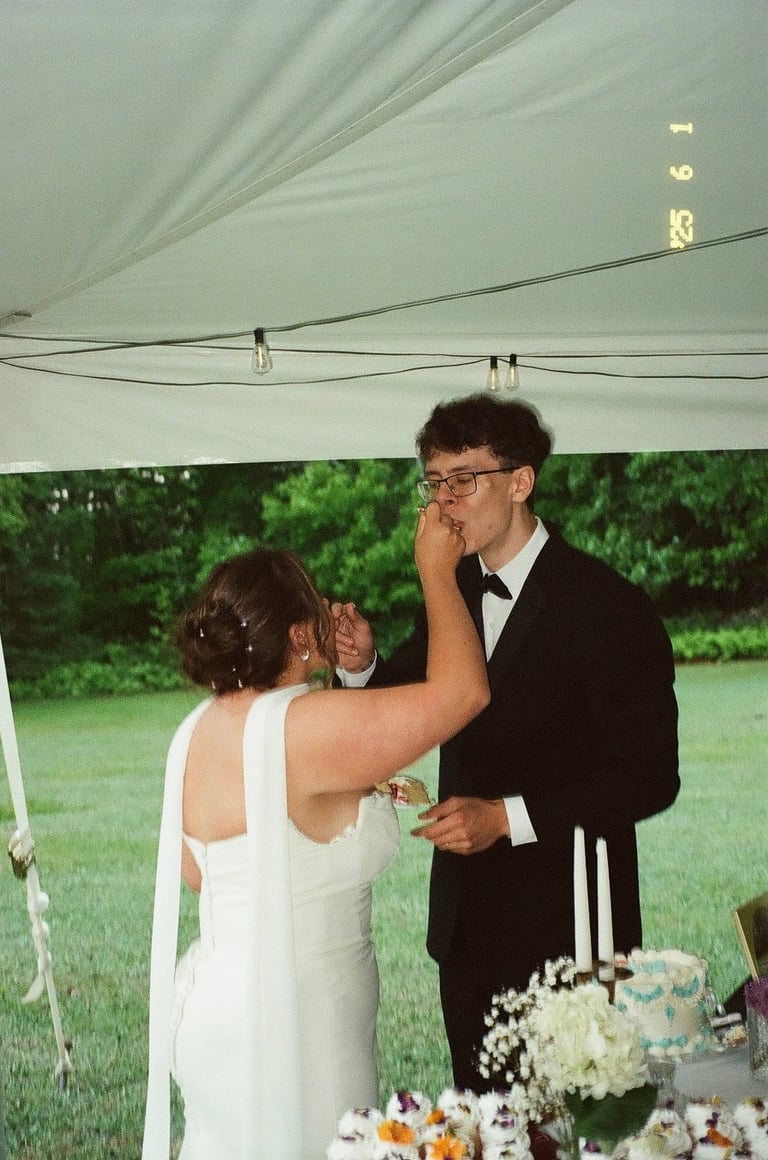 Groom in black tuxedo feeding bride in white dress at outdoor wedding reception under white tent