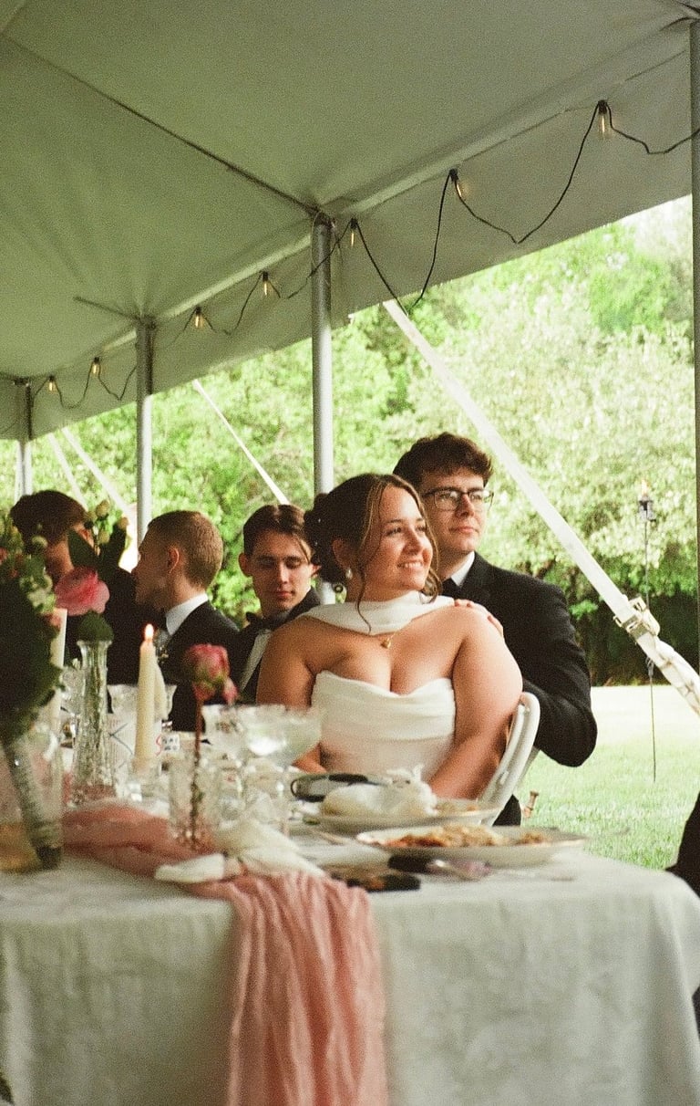 Bride in white strapless dress sitting at table under green tent at outdoor wedding reception with guests