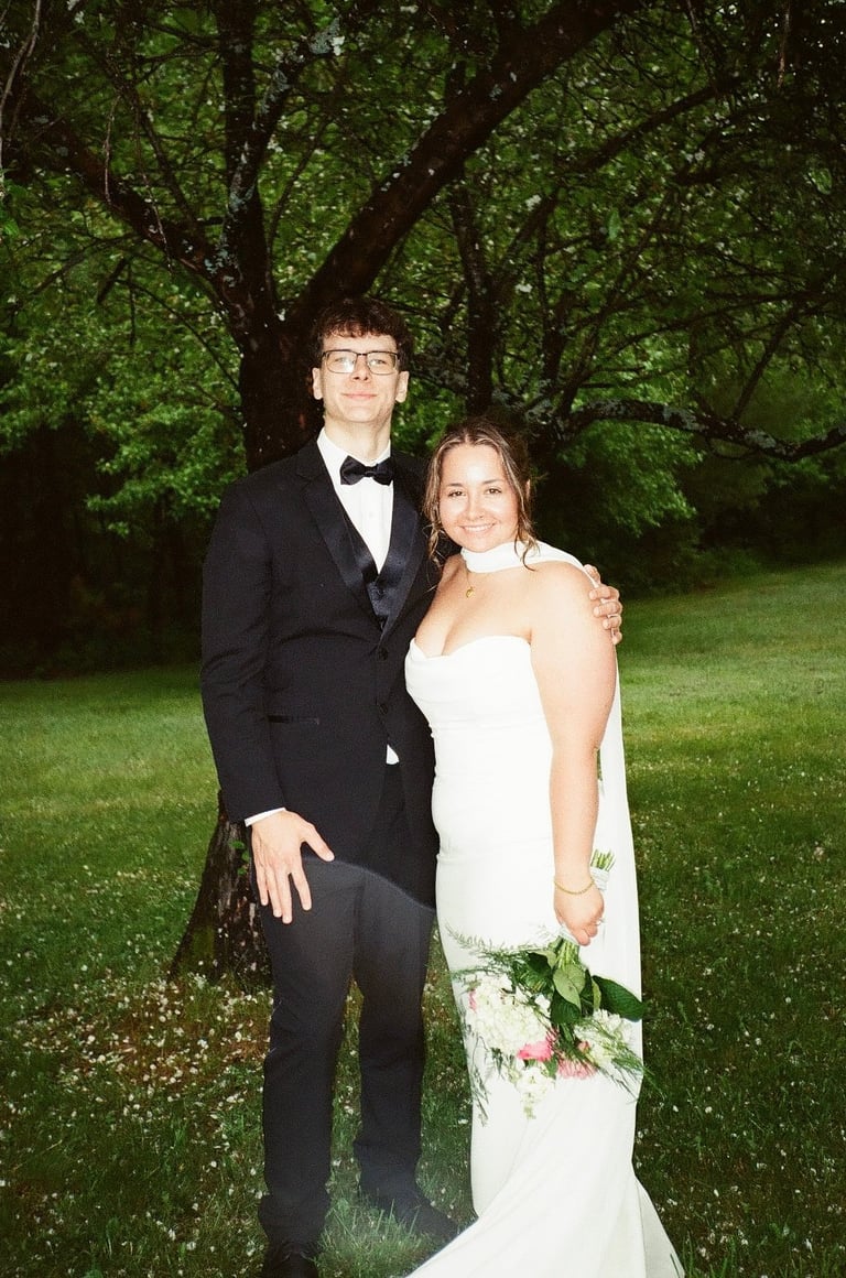 Bride in white gown with bouquet and groom in black tuxedo posing together outdoors under large tree