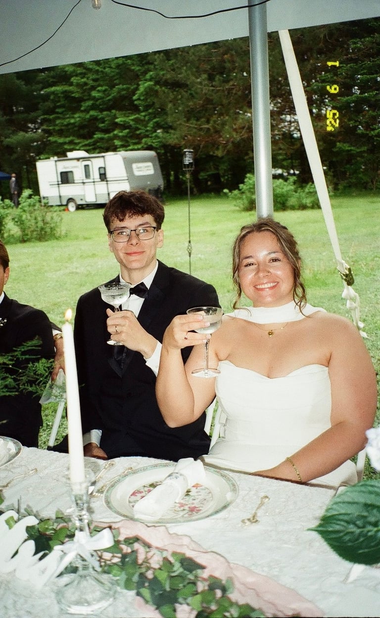 Couple in formal attire toasting at an outdoor wedding reception under a tent with greenery and a caravan in the background